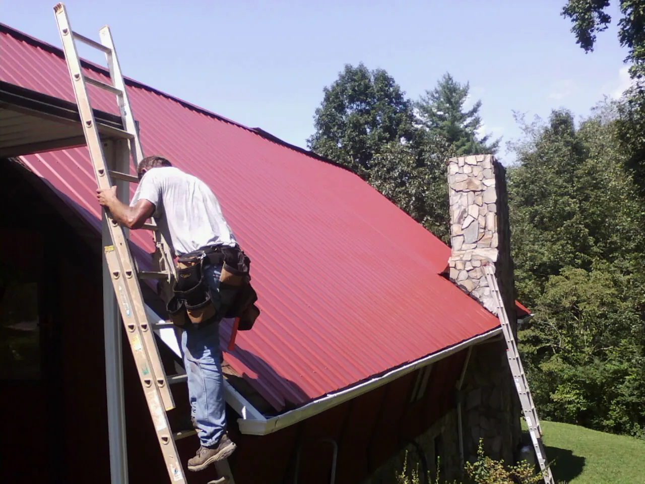 Roofer on a ladder beside a red metal roof and stone chimney — Soffit & Fascia Repair by Roofing Brothers in Jerome